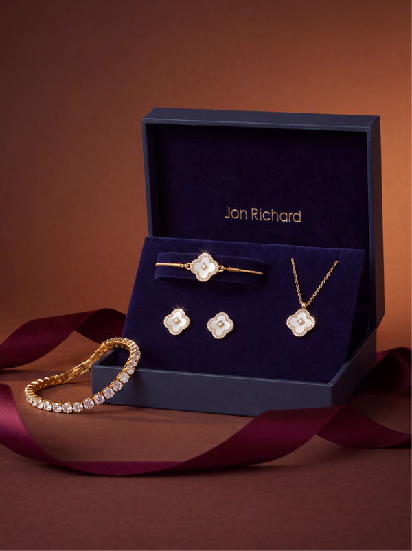 A Jon Richard jewelry set displayed in an open navy presentation box, featuring a gold bracelet, necklace, and earrings with clear stones, alongside a matching gold bracelet on a burgundy ribbon, against a warm brown background.